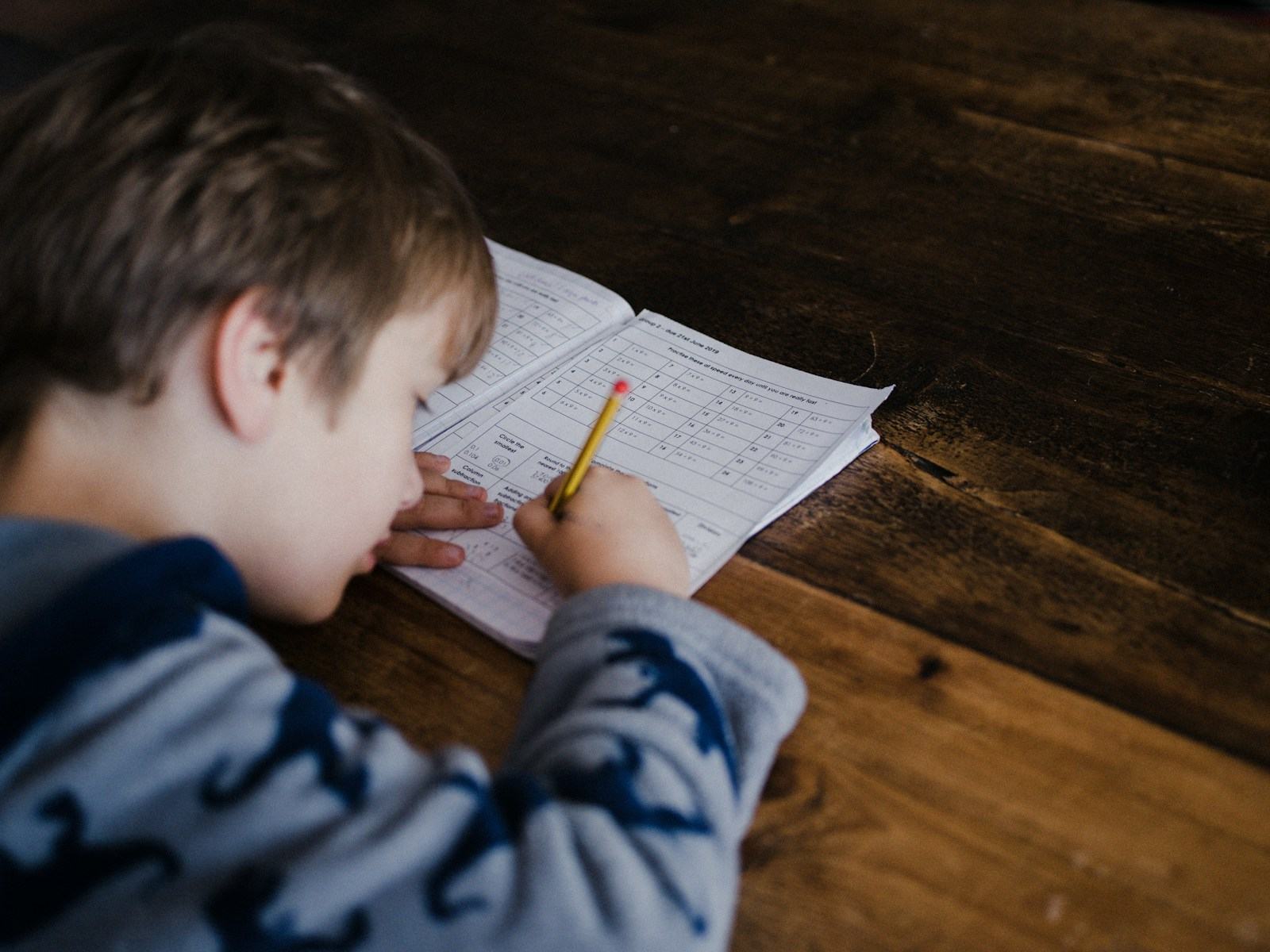 Băiat aplecat peste o fișă de exerciții la matematică, scriind cu creionul la o masă de lemn