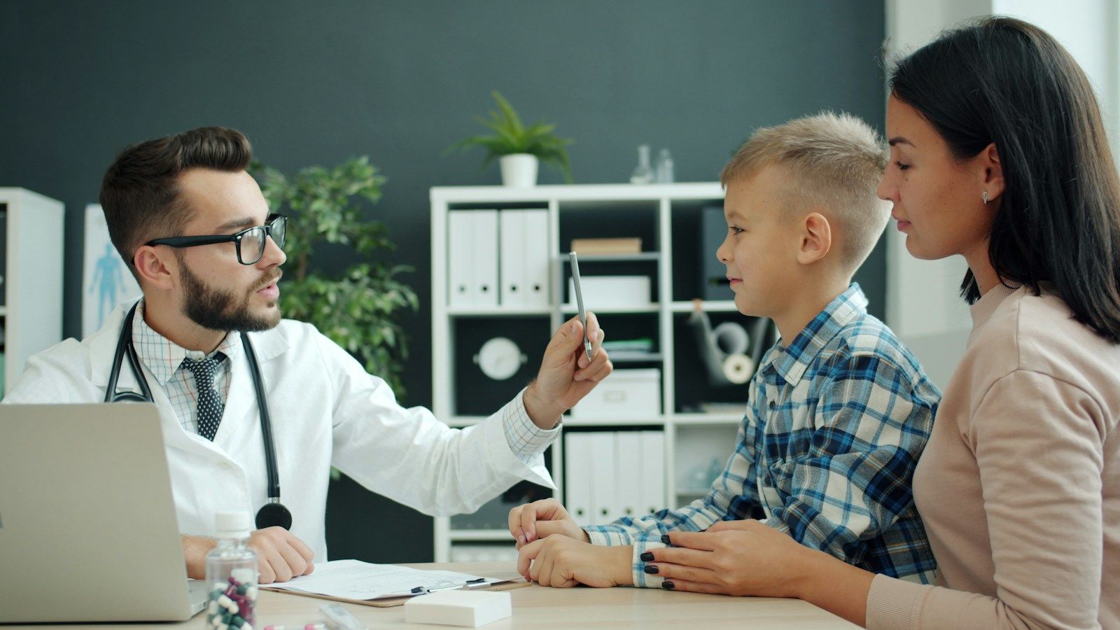 Medic cu ochelari care îi explică ceva unui băiat așezat lângă mama lui, la biroul din cabinet
