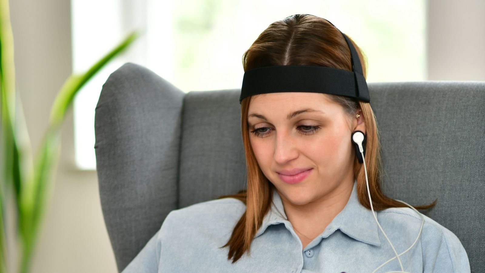 Femeie relaxată într-un fotoliu, cu o bandă cu electrozi pe frunte și un senzor prins de ureche