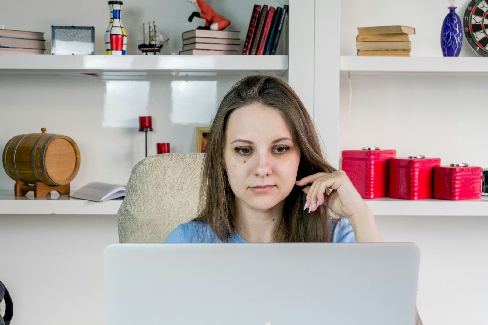 Tânără gânditoare în fața unui laptop, cu mâna la obraz, într-o cameră cu rafturi albe